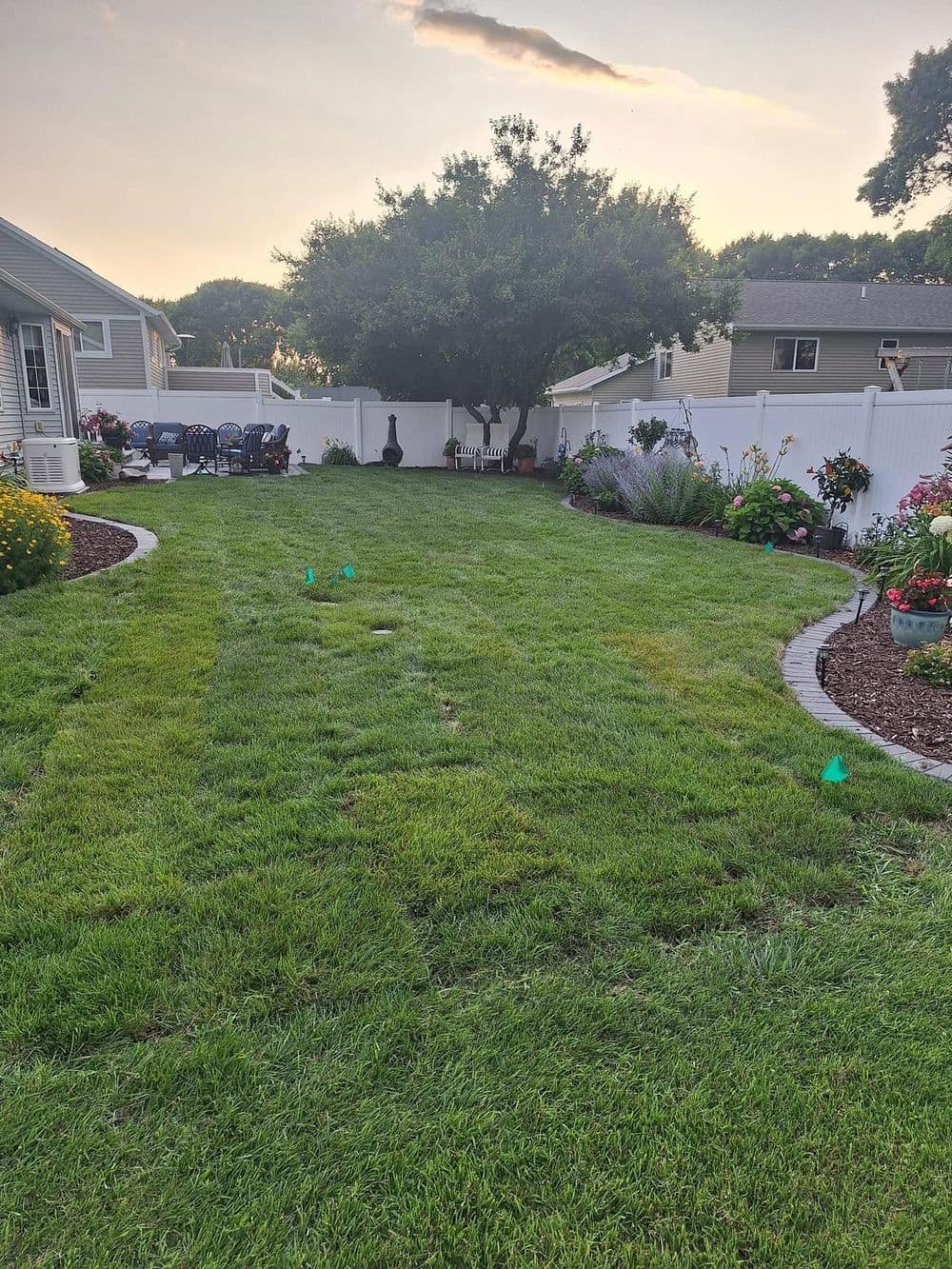 Lush green backyard with flower beds, a tree, and patio furniture under a sunset sky.