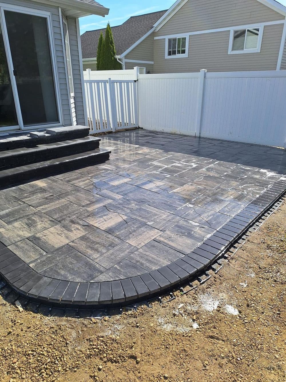 Patio area with patterned paving stones and a black border, surrounded by white fence.