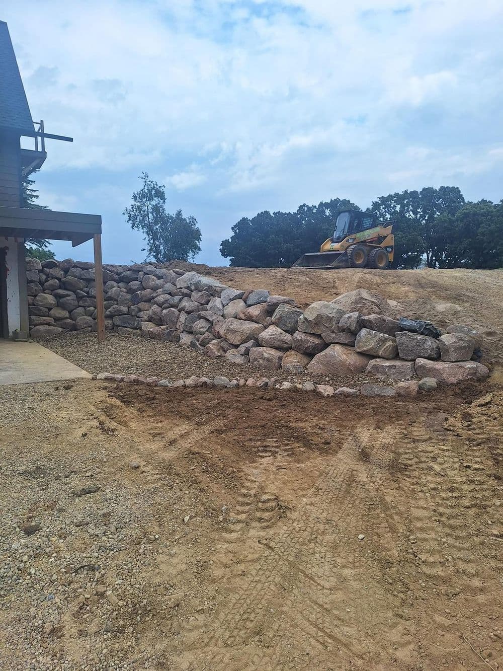Construction site with a skid steer loader and a stone wall under a cloudy sky.
