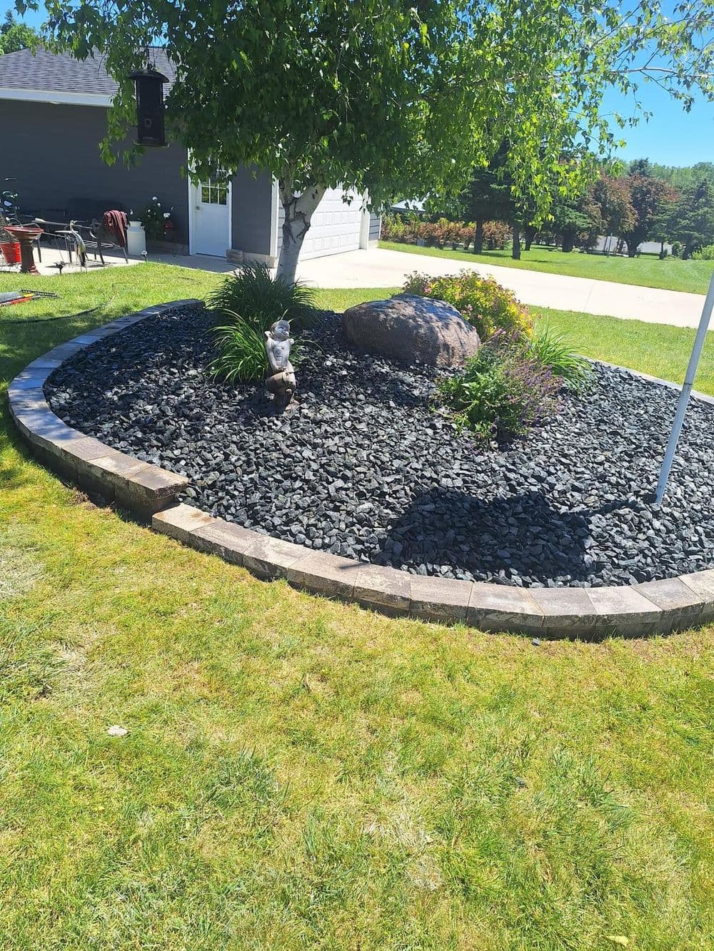 Landscaped garden with black stone, colorful plants, a rock, and a small statue under a tree.