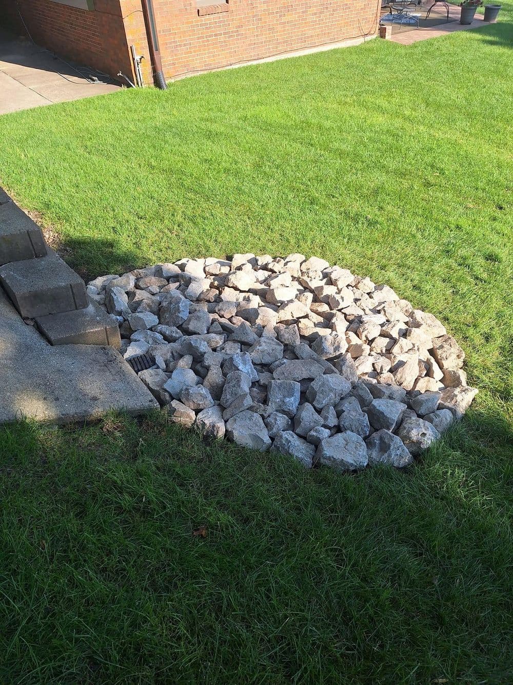 Rock-filled drainage area beside paved steps in a lawn, surrounded by green grass.